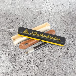 Puede incluir: Un bolígrafo negro y plateado con plumín dorado, sobre una superficie texturizada. El bolígrafo está junto a un estuche de cuero marrón y una caja negra con el texto "Leuchtschreiber" en letras doradas.