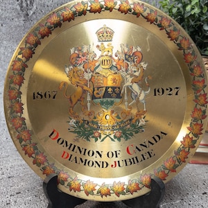 Op de afbeelding: Een goudkleurige, ronde decoratieve schaal met een gedetailleerd embleem en tekst. Het embleem bevat een kroon, leeuwen en een wapenschild. De schaal is omrand met een patroon van herfstbladeren en de tekst "Dominion of Canada Diamond Jubilee."