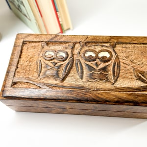 Peut inclure: Une boîte en bois avec un couvercle à charnières, représentant deux hiboux sculptés avec des yeux blancs. La boîte est en bois brun foncé avec un grain de bois naturel.