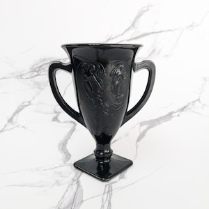 May include: Black glass trophy with two handles and a square base. The trophy features embossed figures on the front and a flared rim. The trophy is displayed on a white marble surface.