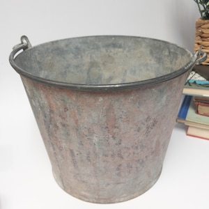 Op de afbeelding: Een verweerde metalen emmer met een taps toelopend ontwerp en een donkere rand. De emmer heeft twee handvatten en een verweerde, gevlekte afwerking met grijze en rode tinten. De emmer staat leeg op een witte ondergrond.