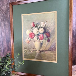 Puede incluir: Una impresión enmarcada de una pintura de bodegón con un jarrón blanco lleno de flores rosas, blancas y rojas. El jarrón está sobre una mesa con un mantel verde. La pintura está enmarcada en un marco de madera marrón con un paspartú verde.