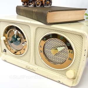May include: Vintage white Silvertone clock radio with two analog clock faces and a radio dial. The radio has a gold trim around the dial and a gold knob on the front. The clock radio is sitting on top of a brown book.