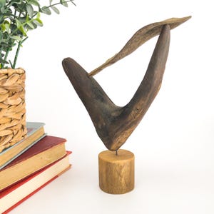 Pode incluir: Escultura de madeira artesanal, apresentando uma forma curva marrom escura com uma peça alongada mais clara que a atravessa. A escultura é montada em uma pequena base cilíndrica de madeira, mostrando o grão natural da madeira. Uma peça única.