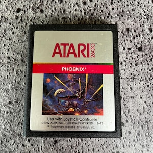May include: A black Atari 2600 game cartridge with a silver label. The label features the Atari logo, the game title "PHOENIX*" in white text, and a colorful image of a spaceship firing lasers. The text "Use with Joystick Controller" is printed below the image, along with copyright information and a trademark notice.