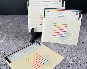 Puede incluir: Tres disquetes de Apple II con rayas de arcoíris de colores. Los disquetes están etiquetados con "Diskware" e incluyen títulos como "Apple Presents... Apple", "DOS 3.3 Sample Programs" y "ProDOS User's Disk".