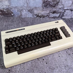 Peut inclure: Un ordinateur personnel Commodore VIC 20 blanc avec un clavier brun et des touches de fonction. Le clavier est en disposition QWERTY standard avec des touches de fonction supplémentaires.