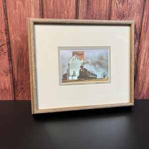 May include: A framed print of a painting depicting a train pulling into a grain elevator. The train is emitting smoke and the scene is set in a rural area. The title of the painting is "A Railroad at Daybreak".