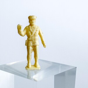 Puede incluir: Una pequeña figura de juguete de soldado de plástico amarillo, con un sombrero y un uniforme. El soldado está de pie sobre una base de acrílico transparente.