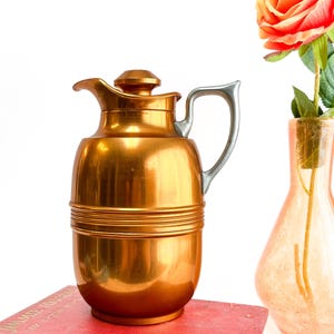 Könnte beinhalten: Eine Vintage-Kanne aus kupferfarbenem Metall mit silberfarbenem Griff und Deckel. Die Kanne ist zylindrisch mit horizontalen Rillen und einem Ausguss. Sie steht auf einem roten Buch. Eine Vase mit einer Rose ist im Hintergrund.
