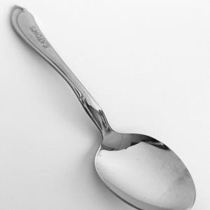 Puede incluir: Una cucharita de plata con un mango adornado y un cuenco redondeado. La cuchara está grabada con la palabra "EATONS".