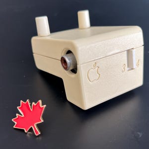 Könnte beinhalten: Ein weißes Apple IIc Disk II Laufwerk mit einem 3,5-Zoll-Diskettenlaufwerksschacht und einem Anschluss für einen externen Monitor. Das Laufwerk hat ein kleines Apple-Logo auf der Vorderseite und einen Schalter mit der Beschriftung "3" und "4".