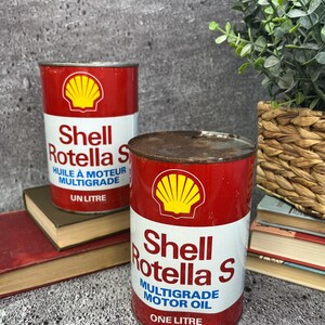 May include: Two vintage cans of Shell Rotella S motor oil. The cans are red with a white Shell logo and white text. The can on the left says "Shell Rotella S Huile à Moteur Multigrade Un Litre". The can on the right says "Shell Rotella S Multigrade Motor Oil One Litre".