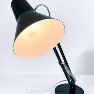 Puede incluir: Una lámpara de escritorio negra con un brazo flexible y una pantalla blanca. La lámpara está encendida y la bombilla es visible.