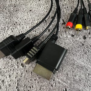 Puede incluir: Un conjunto de cables negros con varios conectores, que incluyen una caja negra con un conector plateado, un cable negro con un conector plateado y tres cables RCA con conectores rojo, amarillo y blanco.
