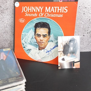 May include: An orange album cover for Johnny Mathis' "Sounds of Christmas" with a portrait of the singer. The album title is in white. A signed photograph and a stack of cassette tapes are also visible.