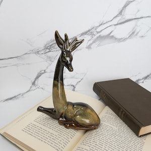 May include: A ceramic deer figurine in brown and green tones, in a reclining pose. The deer has detailed antlers and rests on a book with visible text. The figurine is a decorative item for the home.