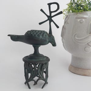 Puede incluir: Una lámpara de aceite de metal verde oscuro con un símbolo Chi Rho en la parte superior, montada sobre un soporte decorativo. La lámpara tiene un pico y un cuerpo texturizado. Al lado, un jarrón blanco de cerámica con forma de cara con vegetación.