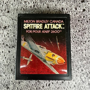 May include: A black Atari 2600 game cartridge with a red, white, and yellow label. The label features a cartoon image of a Spitfire airplane in flight. The text on the label reads "MILTON BRADLEY CANADA SPITFIRE ATTACK FOR/POUR ATARI 2600TM".