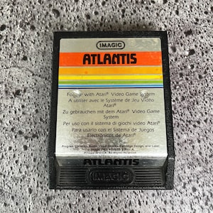 May include: A black Atari video game cartridge with a yellow, orange, and red label. The label reads "IMAGIC ATLANTIS". The back of the cartridge lists instructions for use with the Atari Video Game System in English, French, German, Italian, and Spanish.