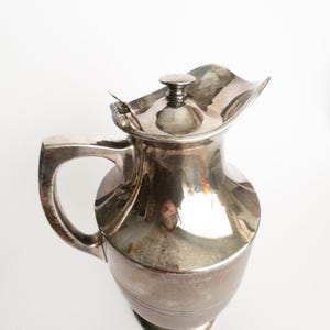 Peut inclure: Une cruche argentée avec une poignée et un couvercle. La cruche a un bec incurvé et une base ronde. Le couvercle est légèrement ondulé. La cruche est en métal et a une surface réfléchissante.