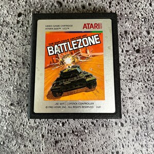 May include: Atari 2600 video game cartridge for Battlezone. The label features a tank in a war scene with the title "BATTLEZONE" in bold white letters. The cartridge is black.