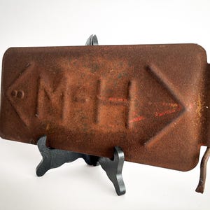 May include: A rusty metal sign with an arrow pointing to the right and the text "MPH" in the centre. The sign is on a black stand.