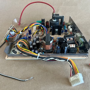 以下が含まれることがあります： コンデンサ、抵抗器、ワイヤーなど、さまざまな電子部品が搭載された回路基板のクローズアップ。基板は金属フレームに取り付けられており、赤いワイヤーが接続されています。基板は、さまざまなワイヤーとコネクターで囲まれています。