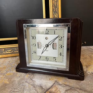 May include: A vintage black Bakelite alarm clock with a silver face and black hands. The clock has a white dial with black numbers and a small second hand dial. The clock is set to 1:55 and the alarm is set to 7:00. The clock is made in England.