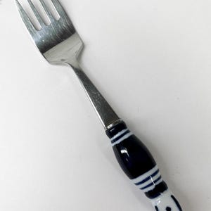 Puede incluir: Un tenedor de plata con un mango de cerámica azul y blanco. El mango tiene un patrón de rayas blancas y un círculo blanco con un punto azul en el centro.