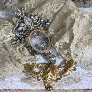 May include: A vintage-style brooch with a large, clear crystal surrounded by smaller rhinestones. The brooch is set on a bed of delicate, ivory lace.