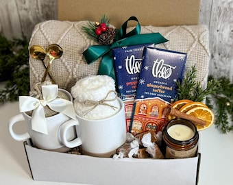 May include: A gift basket filled with cozy items. It includes two white mugs with gold spoons, two chocolate bars, a small candle, a fluffy white item, and wrapped candies. The basket is decorated with a green ribbon and sprigs of greenery.