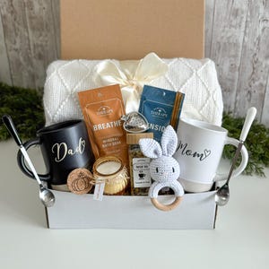 K&ouml;nnte beinhalten: Eine Geschenkbox mit einer wei&szlig;en, strukturierten Decke mit Schleife, einer schwarzen Tasse mit der Aufschrift Dad, einer wei&szlig;en Tasse mit der Aufschrift Mom, zwei Silberl&ouml;ffeln, zwei Badesalzbeuteln, einer Kerze, Kr&auml;utertee und einer grauen H&auml;kelrassel.