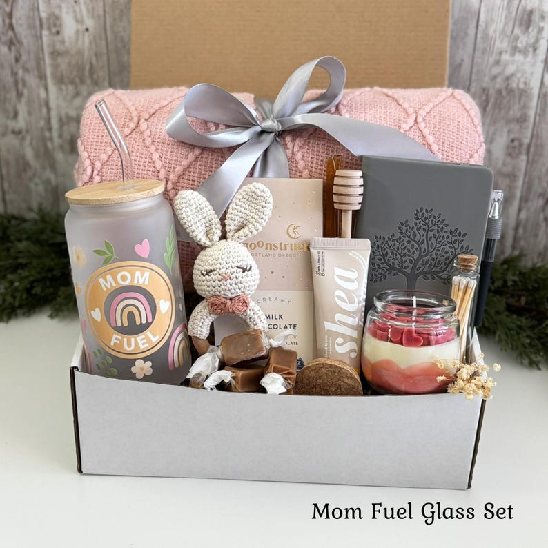 May include: A gift box containing a frosted glass tumbler with a rainbow and Mom Fuel text, a pink knit blanket with a grey ribbon, a crocheted bunny, a chocolate bar, a shea cream tube, a grey journal, a pen, matches, and a candle with heart wax.