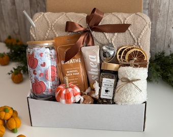 May include: A gift box containing a glass tumbler with a pumpkin print, a metal straw, a bag of mineral bath soak labeled BREATHE, a shea cream tube, a heart-shaped tea infuser, a jar of herbal tea, and a white fuzzy sock with dried orange slices.