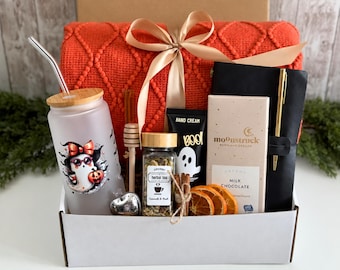 May include: A gift box containing a frosted glass tumbler with a ghost graphic, a wooden honey dipper, a jar of herbal tea, a tube of hand cream, dried orange slices, a Moonstruck milk chocolate bar, a black notebook with a gold pen, and a red blanket.
