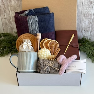 May include: A gift box containing a navy and burgundy plaid throw blanket, a ribbed light blue mug, a brown leather-bound journal with a gold pen, a bubble candle, dried orange slices, cinnamon sticks, a honey dipper, and a pink hair claw.
