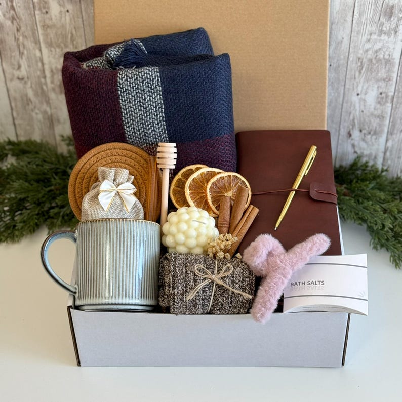 May include: A gift box containing a navy and burgundy plaid throw blanket, a ribbed light blue mug, a brown leather-bound journal with a gold pen, a bubble candle, dried orange slices, cinnamon sticks, a honey dipper, and a pink hair claw.
