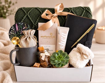 May include: A gift box containing a dark green knit blanket with a beige ribbon, a black notebook with a gold pen, a Moonstruck chocolate bar, a shea cream tube, a gray mug with a gold spoon, a jar of caramels, a succulent, and white fuzzy socks.