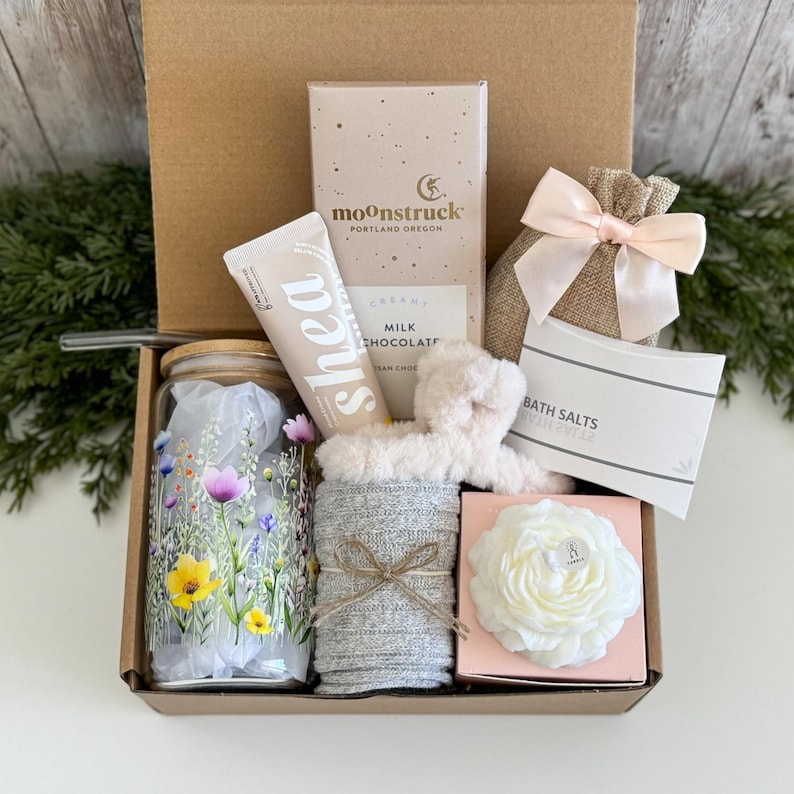 May include: A gift box containing a glass tumbler with a floral print, a tube of shea cream, a Moonstruck milk chocolate bar, a burlap bag with a pink ribbon, a pair of grey socks, a fuzzy headband, and a white rose-shaped candle in a pink box.