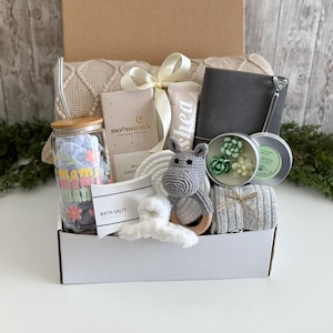 May include: A gift box containing a glass tumbler with a bamboo lid and straw, a Moonstruck chocolate bar, a shea cream tube, a gray notebook, a succulent-shaped candle, a crochet hippo toy, bath salts, a white hair claw, and a gray knit item.