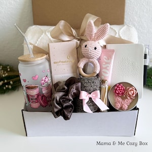 May include: A gift box containing a frosted glass tumbler with a straw, a Moonstruck chocolate bar, a crocheted pink bunny rattle, a rose-scented tube, a white notebook with a tree design, a black pen, a brown scrunchie, and a candle with floral wax.