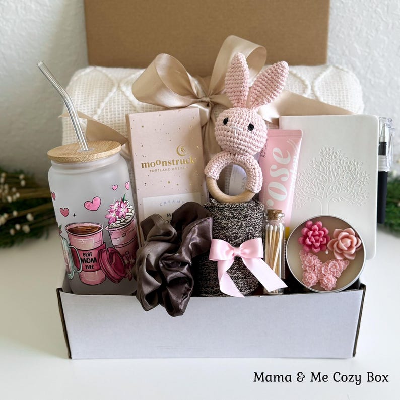 May include: A gift box containing a frosted glass tumbler with a straw, a Moonstruck chocolate bar, a crocheted pink bunny rattle, a rose-scented tube, a white notebook with a tree design, a black pen, a brown scrunchie, and a candle with floral wax.