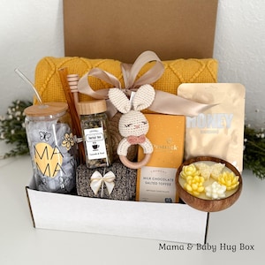 May include: A gift box containing a yellow knit blanket, a glass tumbler with a bamboo lid and straw featuring the text MAMA, a jar of herbal tea, a crochet bunny rattle, a bar of milk chocolate, a honey face mask, and a wooden bowl with flower candles.