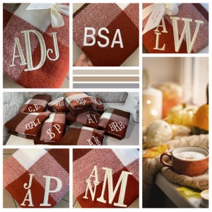 May include: A collection of personalized fleece blankets with various initials embroidered in white on a red and white plaid pattern. The blankets are folded and displayed on a table with a cup of coffee and fall decor in the background.