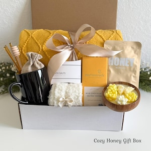 May include: A gift box containing a yellow blanket, a black mug, honey, bath salts, a chocolate bar, and a wooden bowl with decorative flowers. The box is tied with a satin ribbon. The text "Cozy Honey Gift Box" is at the bottom.