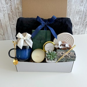 May include: A gift basket filled with items including a blue mug, a candle, a small potted succulent, a notebook, a blanket, and a gold spoon. The basket is lined with brown paper and has a white lid.