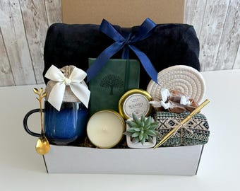 Puede incluir: Una cesta de regalo llena de artículos, incluyendo una taza azul, una vela, una pequeña suculenta en maceta, un cuaderno, una manta y una cuchara dorada. La cesta está forrada con papel marrón y tiene una tapa blanca.