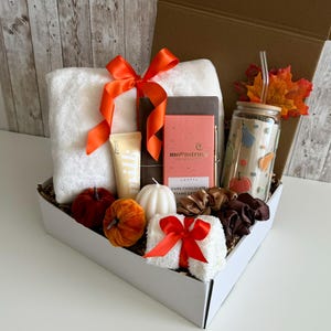 K&ouml;nnte beinhalten: Eine Geschenkbox gef&uuml;llt mit einem wei&szlig;en Handtuch, dunkler Schokolade, einem Glasbecher mit Strohhalm und anderen Artikeln. Die Box ist mit einer orangefarbenen Schleife verziert und enth&auml;lt dekorative K&uuml;rbisse und eine Kerze. Das Thema ist Herbst.
