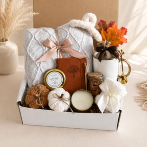 May include: A gift box containing a light gray textured throw blanket with a satin ribbon, a brown notebook with a tree design, a gold-toned pen, a white mug with a gold handle and spoon, a scented candle, two plush pumpkins, and a small jar.